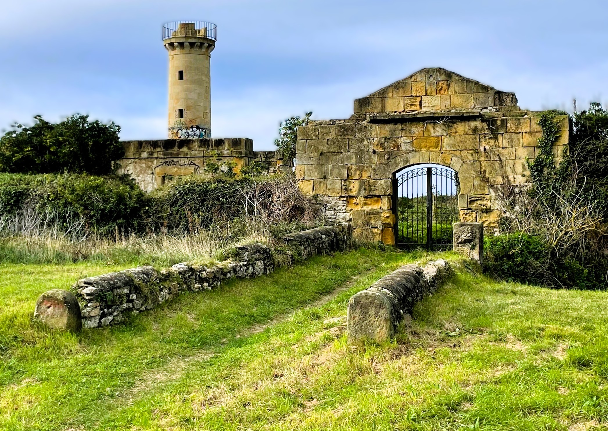 Entrada de piedra con portón de hierro en un prado y una torre de vigilancia antigua al fondo